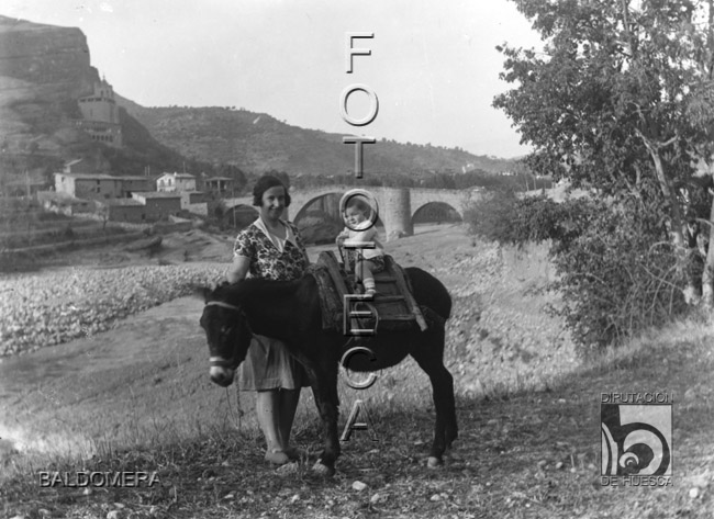 Toñín de Baldomera encima de un burro y su madre a un lado. Al fondo, el puente "de abajo". Antonio López Santolaria. Graus