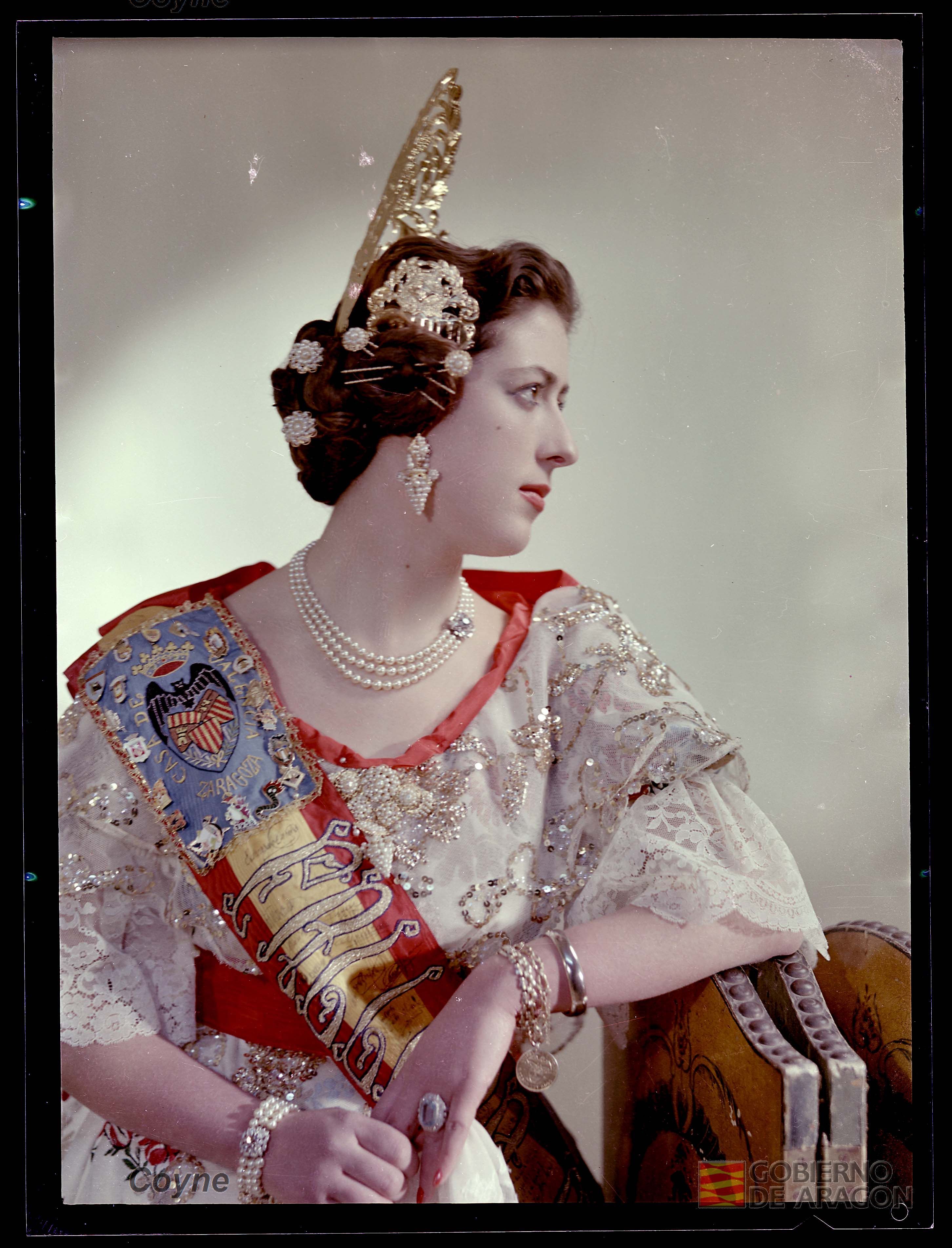 Retrato a color en plano medio de mujer joven seria con vestido, banda de fallera, pendientes, collar, pulseras, anillo y peineta