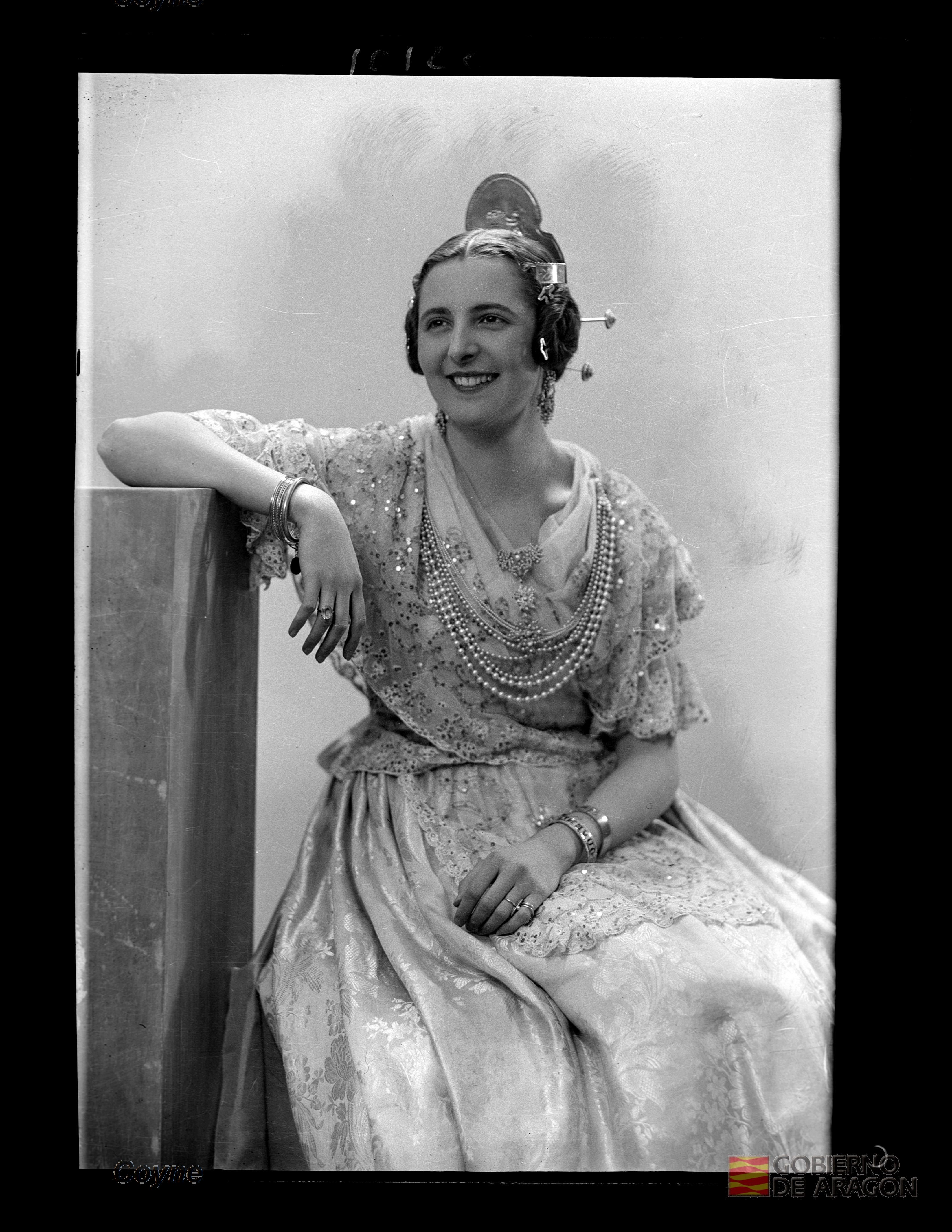 Retrato en plano americano de mujer sonriente de mediana edad con vestido claro, collar, pendientes, peineta y pulseras