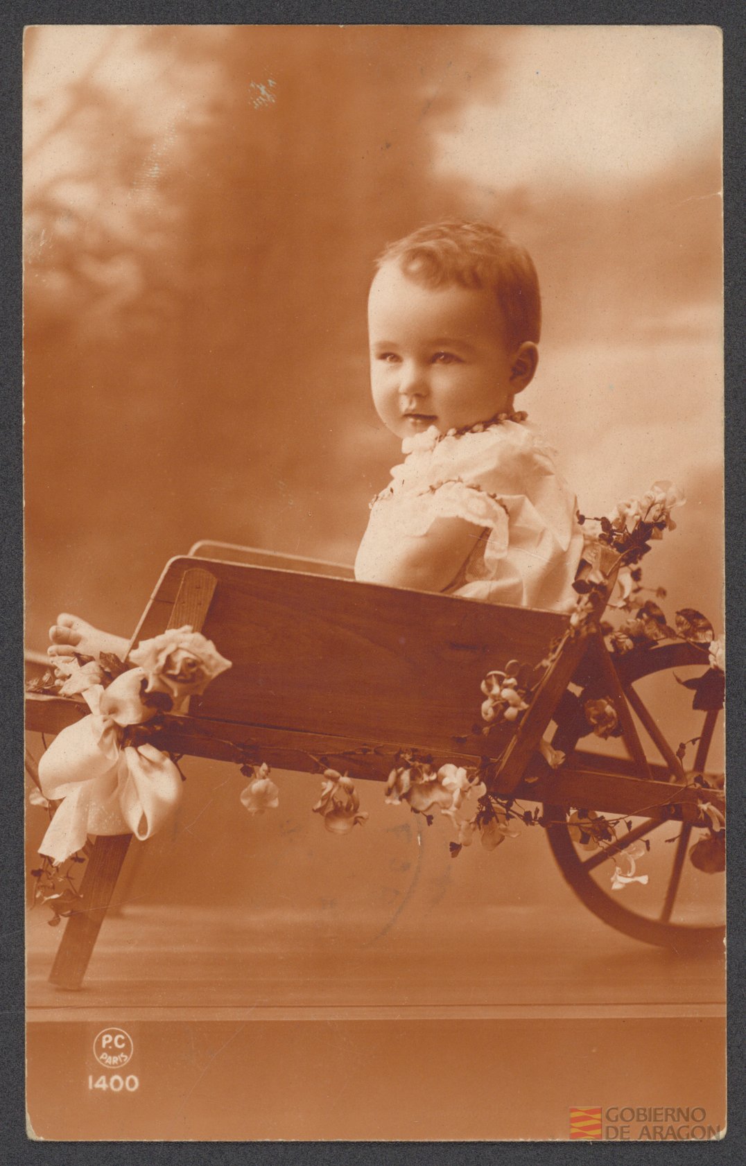 Niño pequeño subido a un carrito de madera adornado con lazos y flores. Editor P-C