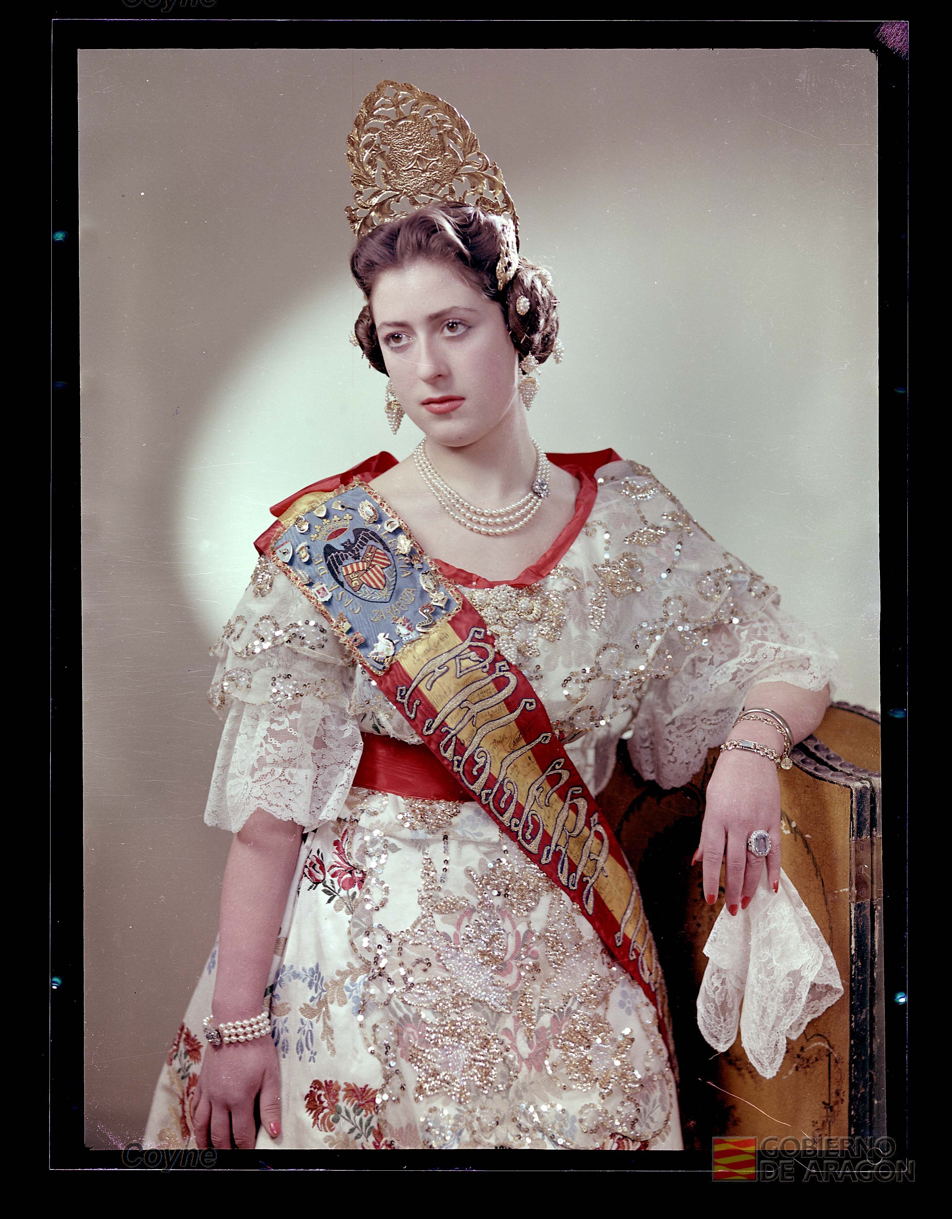 Retrato a color en plano medio largo de mujer joven seria con vestido, banda de fallera, pendientes, collar, pulseras, anillo y peineta