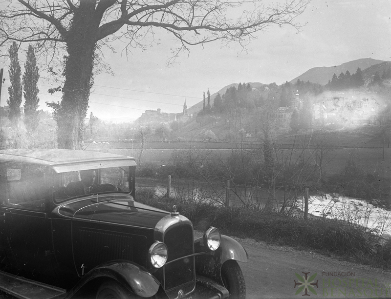 Retrato de hombre conduciendo un coche delante de un pueblo no identificado
